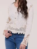 Cardigan corto beige de algodón con bordado en relieve, escote redondo y cierre frontal con botones carey. Mangas largas con detalles de encaje crochet y ruedo con terminación en ondas.