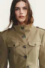 Chaqueta de estilo militar en color beige, con cuello subido, cierre frontal de botones, bolsillos de plastrón con solapa en el delantero y detalle de solapas en el pecho. Presenta mangas largas con hombreras y trabillas decorativas.