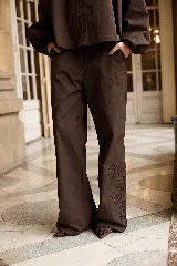 Pantalón de corte recto y tiro medio, color marrón, con detalle de bordado floral en la parte inferior de una de las piernas. Cuenta con cierre y botón frontal.
