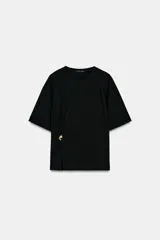 Camiseta de algodón compacto color negro, con cuello redondo, manga corta y detalle de aplique metálico en el frente que genera un ligero fruncido lateral.