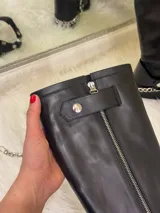 Bota alta tipo biker de cuero negro con cierre metálico lateral que recorre toda la caña. Presenta un diseño versátil con estribo y cadena metálica desmontables en la zona del tobillo, taco cuadrado y plataforma delantera.