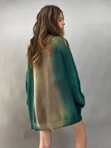 Buzo tejido de lana merino con diseño oversize, manga ranglan y cuello levemente elevado. Presenta un efecto de teñido degradado que combina tonos verdes y beige.