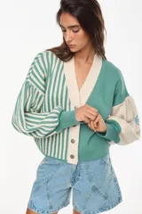 Cardigan tejido bicolor en tonos verde y crudo, con diseño de rayas verticales en la mitad derecha y liso en la izquierda. Presenta cuello en V, cierre frontal con botones y mangas abullonadas con puños ajustados.