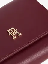 Billetera trifold de color borgoña con acabado liso y cierre de solapa. Presenta una insignia metálica dorada con el monograma TH en el frente.