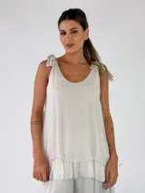 Top musculosa color beige, con escote redondo y tirantes ajustables con lazos en los hombros. Presenta un diseño holgado con detalle de volado en el ruedo.