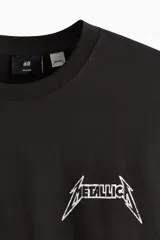 Remera negra de corte holgado con hombros caídos y cuello redondo. Presenta un estampado frontal pequeño con el logo de Metallica y la inscripción 'U.S. Tour 1985'.