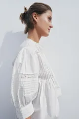 Blusa de algodón color blanco con cuello redondo y mangas abullonadas. Presenta detalles de puntilla a tono en el pecho y las mangas, bajo recto y cierre posterior con abertura y botón.