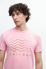 Remera de algodón con efecto desgastado o acid wash, color rojo coral. Presenta un estampado frontal con líneas onduladas en azul marino y el texto "TROPIC TRIBE CREW" y "UNIFORM".