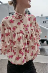 Blusa de cuello subido y manga larga con estampado floral en tonos rosados. Presenta un diseño con volantes en el pecho y a lo largo de las mangas, con cierre posterior mediante botones ocultos.