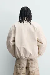 Cazadora de algodón color beige con efecto lavado, corte relaxed fit, cuello solapa y cierre frontal con cremallera. Cuenta con bolsillos frontales tipo canguro, puños y bajo elásticos, y aplicaciones de parches bordados a contraste en el delantero.
