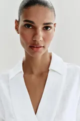Blusa blanca confeccionada en lino, con cuello polo, escote en V y mangas largas amplias. Presenta un diseño holgado y bajo asimétrico.
