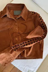 Camisa marrón de gamuza con cuello clásico, cierre con botones a presión y mangas largas con detalle de calados artesanales.