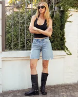 Bota de caña alta confeccionada en cuero negro, con suela track de goma. Presenta un diseño versátil con dos hebillas ajustables en la parte superior de la caña y una correa decorativa con tachas metálicas y argollas en la zona del tobillo.