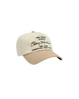 Gorra de cinco paneles con corona en color crema y visera en color tostado. Presenta un diseño bordado en la parte frontal con el logo y texto de la marca en color negro.