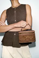 Bolso formato vanity de gamuza color marrón, con asa de mano superior y correa bandolera extraíble y ajustable. Presenta cierre de doble cremallera metálica dorada y detalles de pespuntes en el cuerpo.