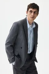 Blazer cruzado de color gris medio, con solapas de pico, ojal decorativo y cierre de doble botonadura. Presenta bolsillo superior insertado, bolsillos delanteros ribeteados con solapa y botones decorativos en los puños.