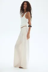 Pantalón de tejido liso color blanco roto o crema, de corte ancho y tiro alto con cintura elástica fruncida. Presenta pliegues frontales que le dan volumen a la pierna.