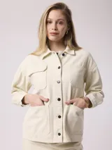 Campera de jean color beige, de corte recto, con cuello clásico, cierre frontal con botones metálicos, bolsillos de parche en el pecho y mangas largas con puños abotonados.