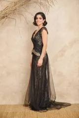 Vestido largo de tul y encaje negro con detalles dorados en los hombros y la cintura. Presenta escote en V cruzado, corte ajustado y cola de tul.
