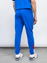 Pantalón de jogging azul de felpa, con detalles de piping y franjas laterales en rojo y blanco. Incluye el escudo del Club Nacional de Football en el muslo derecho.