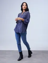 Blusa de gasa con detalles de encaje crochet en hombros y espalda. Presenta cuello a la base con escote en V, cierre frontal con botones y mangas tres cuartos amplias con puño abotonado. El diseño cuenta con un corte holgado y ruedo irregular, siendo más largo en la parte posterior.