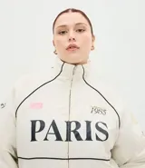 Campera bomber de microfibra color beige con estampado frontal de lettering 'PARIS 1985'. Presenta cierre frontal con cremallera, cuello alto, bolsillos funcionales y puños y ruedo ajustados con elástico.