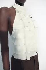 Top de cuello halter color blanco, con múltiples volantes en cascada en todo el frente y la espalda descubierta con cierre de botones y lazada en el cuello.