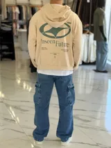 Hoodie de corte boxy fit color beige, con capucha y estampa gráfica verde en la espalda con la inscripción "Unseen Future" y detalles tipográficos.