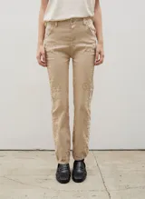 Pantalón de denim color beige con diseño de tachas metálicas distribuidas a lo largo de las piernas. Presenta un corte ajustado, cierre frontal con botón y bolsillos laterales.
