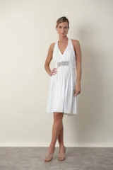 Vestido corto de color blanco con escote halter y espalda descubierta. Presenta un diseño drapeado en el busto y una banda en la cintura decorada con bordados y pedrería. La falda tiene un corte fluido con movimiento.