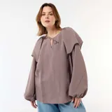 Blusa fluida de manga larga con diseño a rayas en tonos marrón y blanco, con escote redondo adornado con una pequeña abertura y tiras para anudar, y un gran volante superpuesto en los hombros.