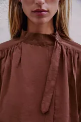 Camisa bordó de cuello alto ajustable con doble hebilla y cierre frontal con botones ocultos por solapa. Tiene mangas cortas con trabillas y diseño holgado.