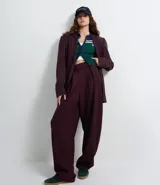 Pantalón de sastrería corte barrel, color marrón oscuro, con pliegues delanteros, cintura con cierre desplazado y piernas anchas.