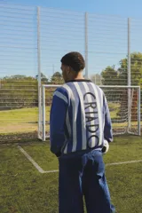 Buzo de punto estilo retro inspirado en la camiseta de la selección de 1930, con diseño de rayas verticales en azul y blanco. Presenta cuello polo negro, puños a contratono y la palabra 'CRIADO' bordada en letras grandes sobre la espalda.