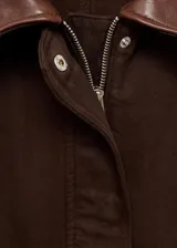 Chaqueta corta de algodón en color marrón oscuro, con cuello de efecto piel en tono contrastante. Presenta cierre frontal con cremallera y botones a presión, además de dos bolsillos frontales con solapa y cierre de botón.