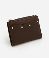 Billetera mediana color marrón, confeccionada en poliuretano con textura lisa y detalle de gamuza en la parte superior. Presenta cierre superior con cremallera dorada y aplicación de tachas metálicas decorativas en el frente.
