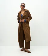 Trench coat largo de gamuza sintética color marrón oscuro, con cuello solapa, cierre frontal cruzado con botones y cinturón ajustable en la cintura. Presenta mangas largas con reguladores en los puños y forro interior.