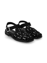 Sandalia estilo cangrejera color negro, confeccionada en cuero gamuza, con tiras entrelazadas decoradas con tachas metálicas plateadas. Cuenta con tira ajustable en el talón con hebilla y suela plana de caucho.