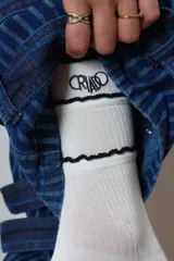 Medias blancas de caña media con tejido acanalado, detalles de volados negros en el puño y bordado con la palabra "Criado" en color negro.