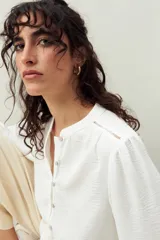 Camisa color marrón claro con cuello mao, mangas 3/4 abullonadas y cierre frontal con botones. Presenta pequeños pliegues en los hombros y detalle calado sutil.