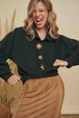 Camisa de pana fina color verde oscuro, de manga larga y corte holgado. Presenta cuello clásico y cierre frontal con botones, decorada con tres prendedores metálicos dorados en forma de sol, flor y estrella de mar.