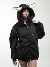 Campera negra con cierre frontal, capucha con orejas decorativas blancas con tachas, y detalles de tachas metálicas en el frente y hombros. Diseño de corte regular.