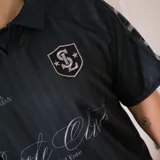 Camiseta de fútbol estilo boxy de manga corta y cuello polo, en color negro con sutiles rayas verticales. Presenta inscripciones estampadas y escudo bordado en el pecho.