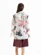 Camisa de seda color blanco con estampado floral y de dragones en tonos rojos, azules y verdes. Tiene cuello clásico, mangas largas con puños ajustables y botones forrados.