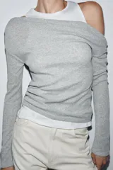 Camiseta de manga larga gris melange con cuello redondo y diseño asimétrico que simula una doble camiseta, con una capa inferior blanca visible en el cuello y el bajo.