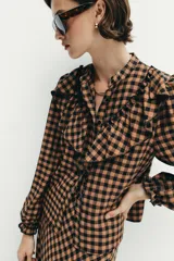 Blusa de manga larga con estampado de cuadros vichy en tonos marrón y negro. Presenta cuello redondo, cierre frontal con botones y detalle de volantes en el pecho.