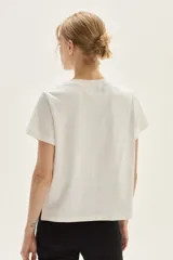 Remera blanca de algodón con cuello redondo y manga corta. Presenta un estampado bordado central con motivos florales y un animal (posiblemente un conejo o liebre) en tonos lila, morado y marrón.