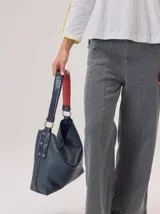 Cartera tipo hobo de cuero azul con textura granulada. Presenta un diseño holgado, con una correa de hombro bicolor que combina azul y rojo, sujeta por argollas metálicas. Incluye una placa metálica pequeña con el logo de la marca en la parte inferior frontal.