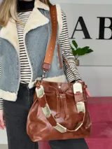 Bolso tipo bucket bag Chabas modelo 3.1, confeccionado en cuero genuino de color marrón. Cuenta con cierre de cordón ajustable, dos tipos de asas (una corta de cuero con detalles decorativos y otra larga tipo bandolera con tira textil) y diseño sin forro interior.