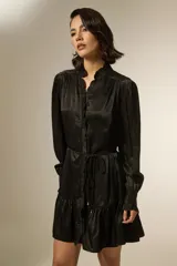 Vestido corto de satén negro, con cuello mao y detalle de volados en la botonadura frontal. Posee mangas largas con puños ajustados y un cordón fino en la cintura para entallar, terminado con pequeñas cuentas doradas.